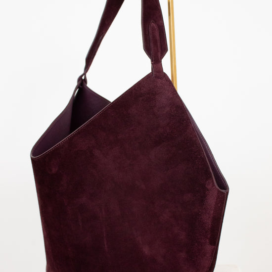 A large slouchy shoulder bag in deep burgundy suede, displayed on a minimalist gold metal stand against a clean white background.
