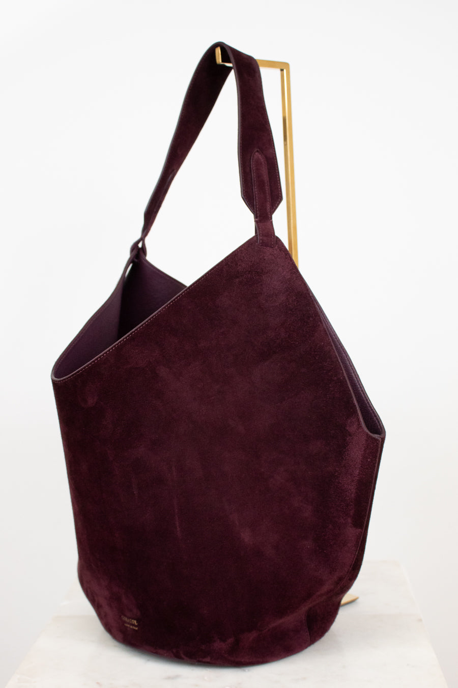 A large slouchy shoulder bag in deep burgundy suede, displayed on a minimalist gold metal stand against a clean white background.