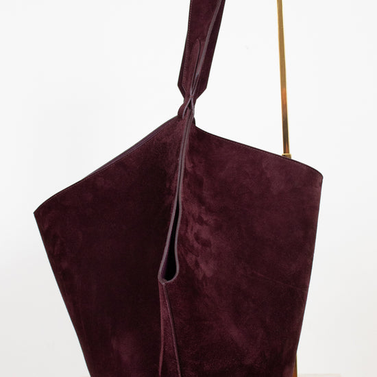 A large, slouchy suede shoulder bag in deep burgundy, displayed on a minimalist gold metal stand against a clean white background.