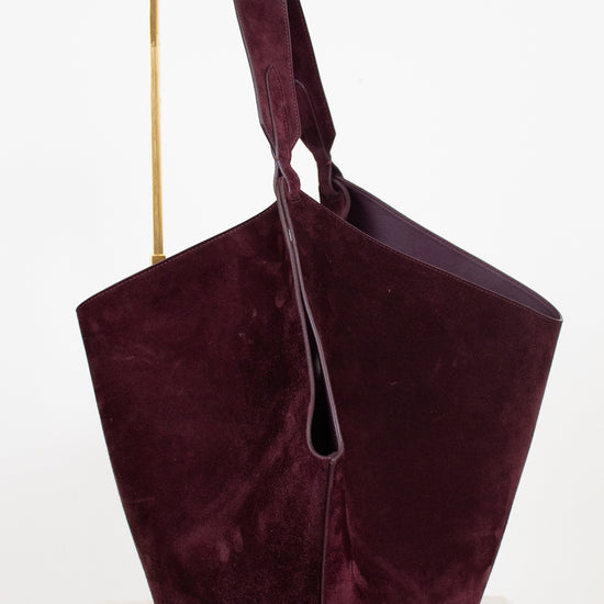 A large slouchy shoulder bag in deep burgundy suede, displayed on a minimalist gold metal stand against a clean white background.