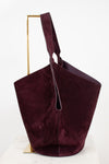 A large slouchy shoulder bag in deep burgundy suede, displayed on a minimalist gold metal stand against a clean white background.
