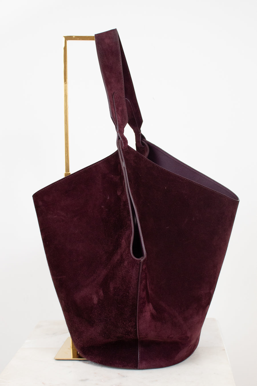 A large slouchy shoulder bag in deep burgundy suede, displayed on a minimalist gold metal stand against a clean white background.