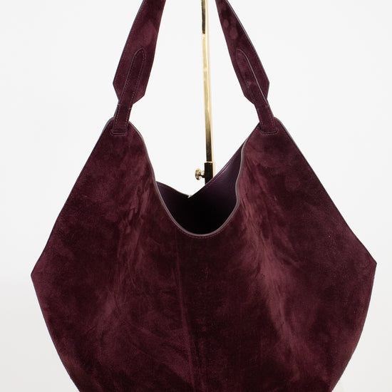 A large slouchy hobo shoulder bag in deep burgundy suede, displayed on a white surface against a white background with a gold-colored metal stand visible behind it.