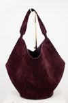 A large slouchy hobo shoulder bag in deep burgundy suede, displayed on a white surface against a white background with a gold-colored metal stand visible behind it.
