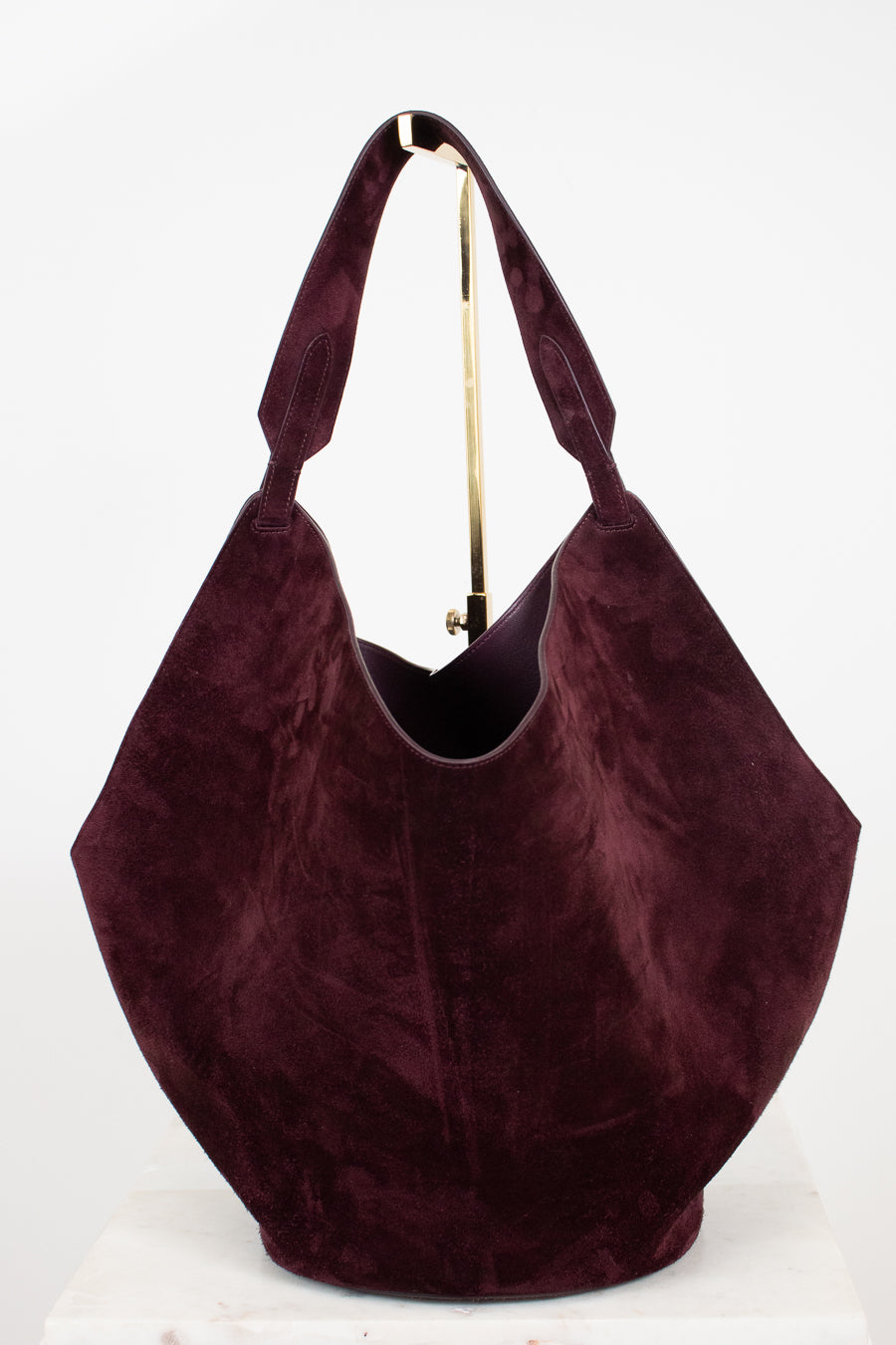 A large slouchy hobo shoulder bag in deep burgundy suede, displayed on a white surface against a white background with a gold-colored metal stand visible behind it.