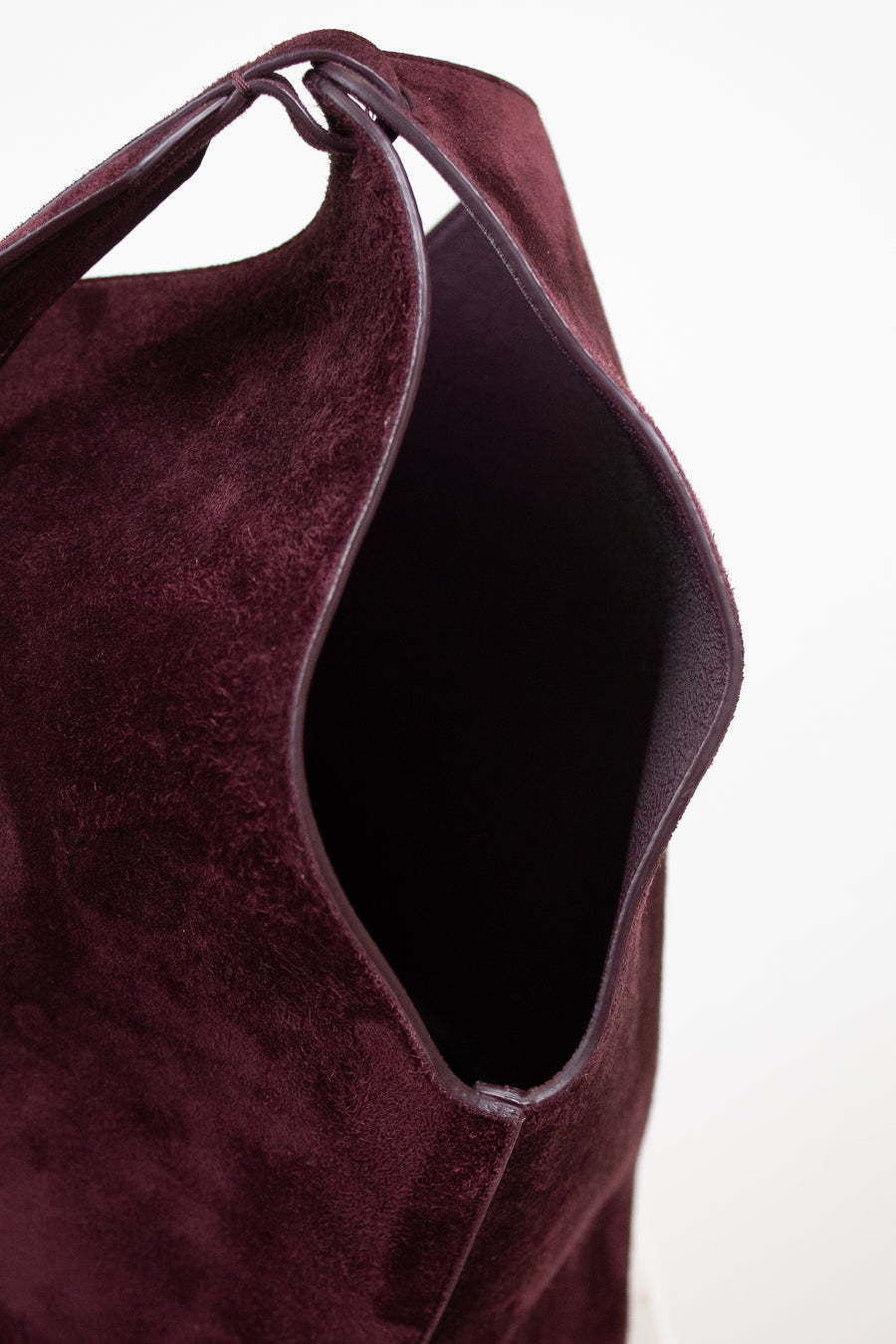 Close-up of a deep burgundy suede shoulder bag, showing the texture of the material and the dark interior, set against a plain white background.