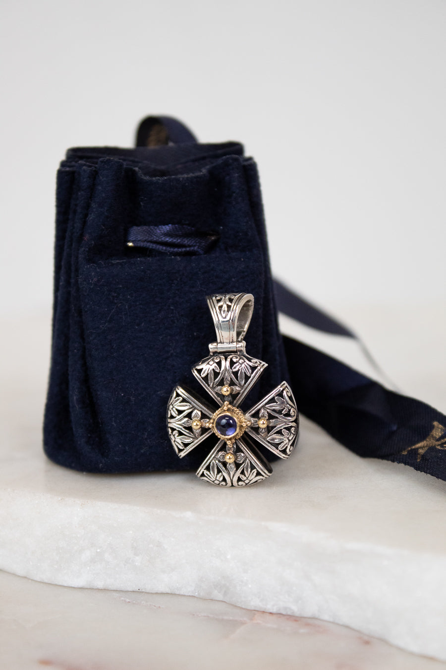Silver and gold cross pendant leaning on navy dust bag on white marble background