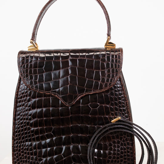 A dark brown alligator leather handbag with a top handle and a separate coiled shoulder strap, displayed against a clean white background.