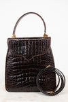A dark brown alligator leather handbag with a top handle and a separate coiled shoulder strap, displayed against a clean white background.