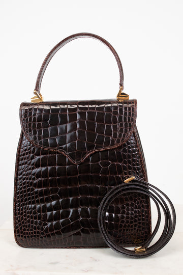A dark brown alligator leather handbag with a top handle and a separate coiled shoulder strap, displayed against a clean white background.