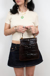 A model wearing a cream short-sleeve crop top, a dark denim mini skirt, and multiple gold necklaces and rings, holding a dark brown alligator leather handbag against a plain light gray background.