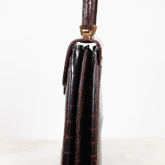 A side profile view of a dark brown alligator leather clutch with a top handle, resting on a light marble surface against a white background.