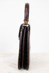 A side profile view of a dark brown alligator leather clutch with a top handle, resting on a light marble surface against a white background.