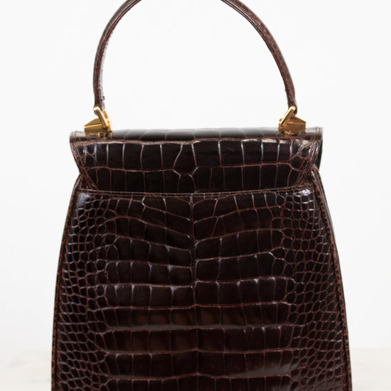 A dark brown alligator leather handbag with a top handle and gold-tone hardware, photographed against a plain white background.