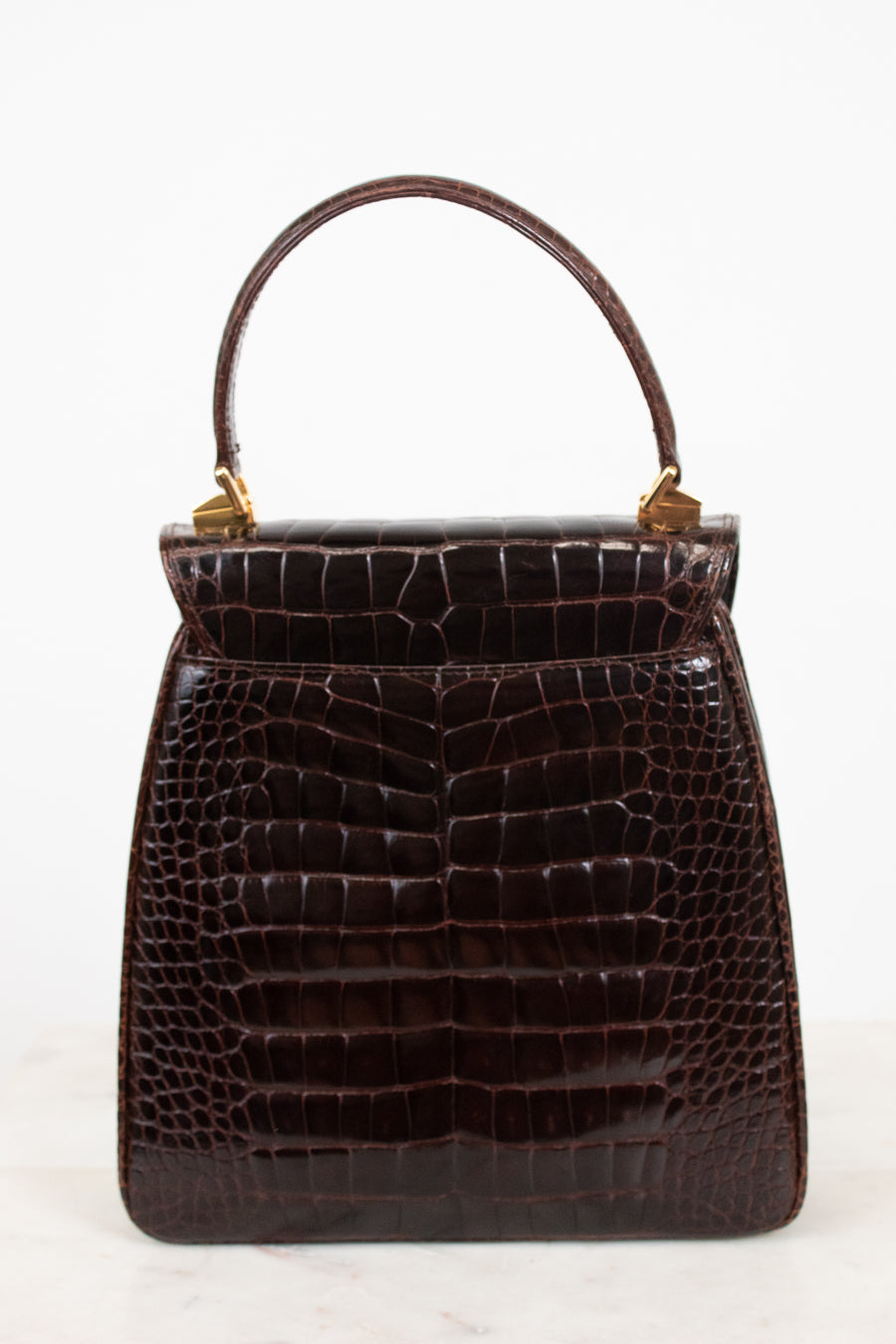 A dark brown alligator leather handbag with a top handle and gold-tone hardware, photographed against a plain white background.