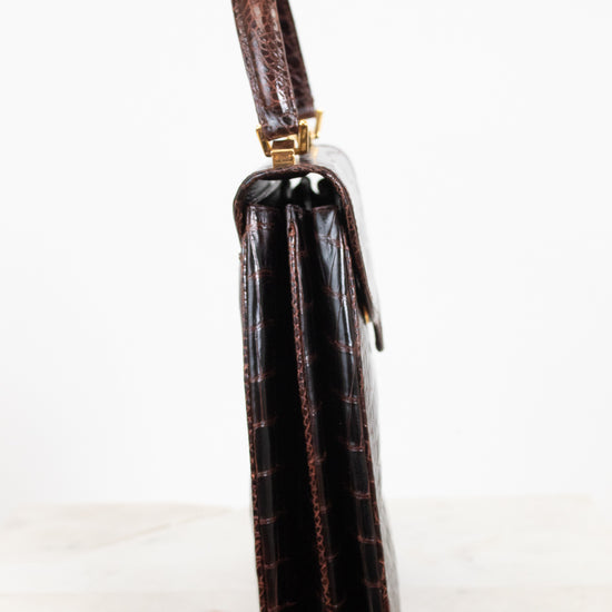 A side profile view of a dark brown alligator leather handbag with gold-tone hardware, standing on a light marble surface against a white background.