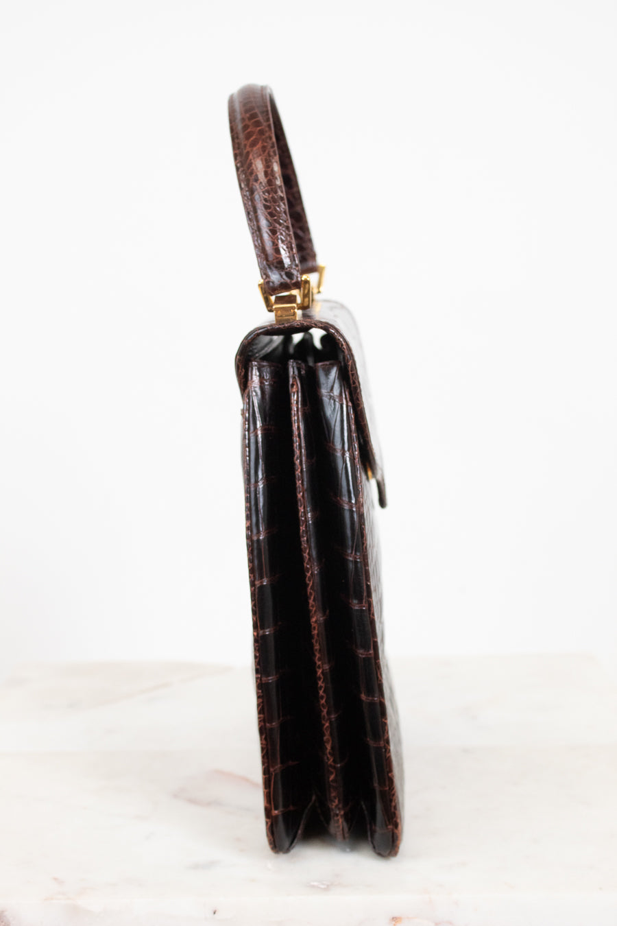 A side profile view of a dark brown alligator leather handbag with gold-tone hardware, standing on a light marble surface against a white background.