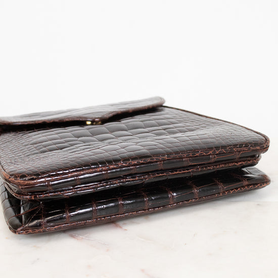 A dark brown alligator leather bag with a fold-over closure flap, resting on a white marble surface against a white background.