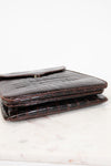 A dark brown alligator leather bag with a fold-over closure flap, resting on a white marble surface against a white background.