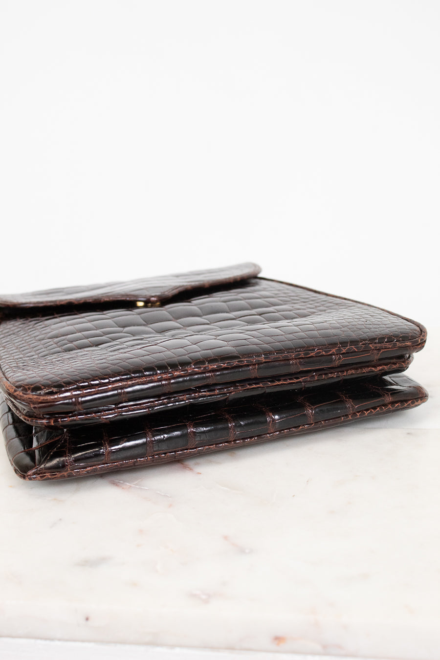 A dark brown alligator leather bag with a fold-over closure flap, resting on a white marble surface against a white background.