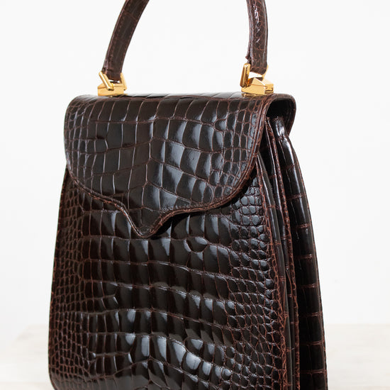 A dark brown alligator leather handbag with a top handle and gold-tone hardware, shown on a light gray background.