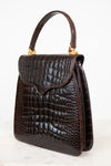 A dark brown alligator leather handbag with a top handle and gold-tone hardware, shown on a light gray background.