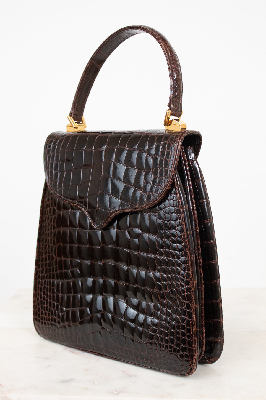 A dark brown alligator leather handbag with a top handle and gold-tone hardware, shown on a light gray background.