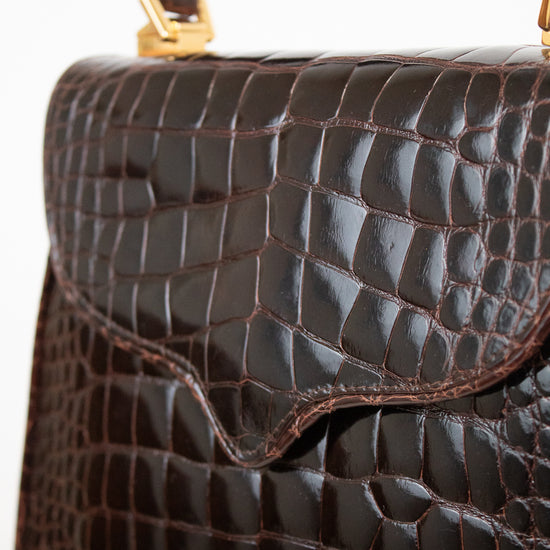 Close-up of a dark brown alligator leather handbag with gold-tone hardware and a top handle against a plain white background.