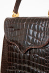 Close-up of a dark brown alligator leather handbag with gold-tone hardware and a top handle against a plain white background.