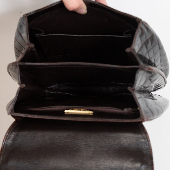 A close-up of a dark brown alligator leather handbag being opened by a hand, showing its interior compartments and a small gold clasp.