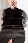 A close-up of a dark brown alligator leather handbag being opened by a hand, showing its interior compartments and a small gold clasp.