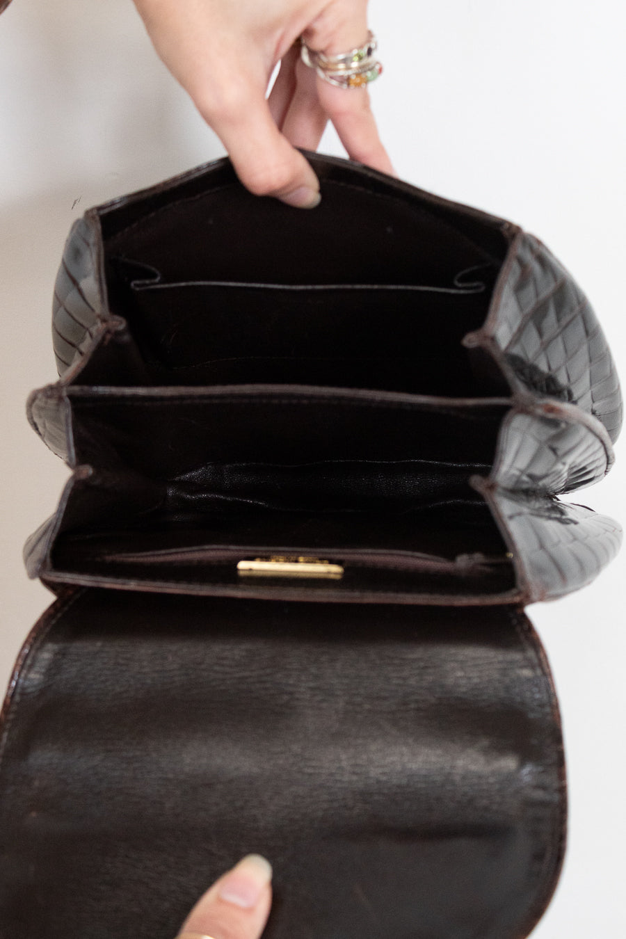 A close-up of a dark brown alligator leather handbag being opened by a hand, showing its interior compartments and a small gold clasp.
