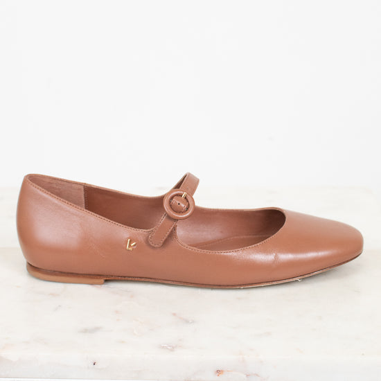 Brown leather flat shoe with a round buckle on a light marble surface.