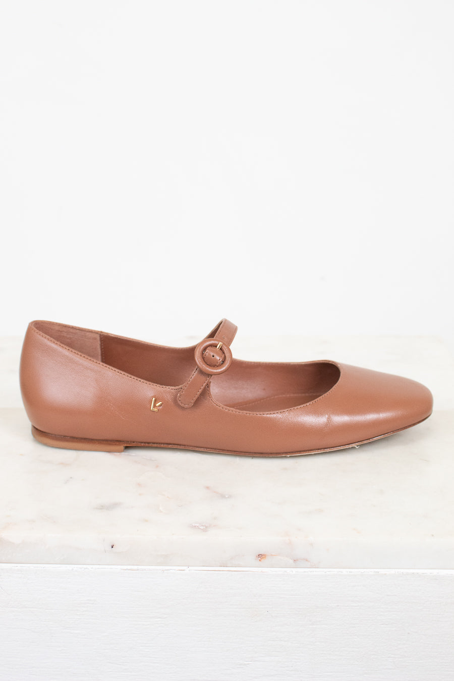 Brown leather flat shoe with a round buckle on a light marble surface.