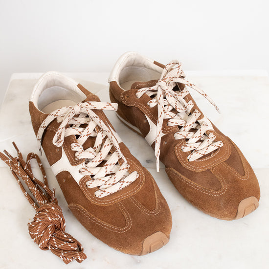 A pair of brown suede sneakers with cream and brown patterned laces, displayed on a white marble surface against a white background.