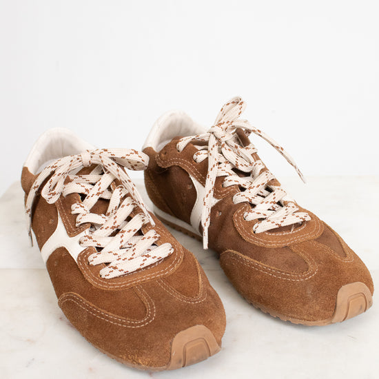 A pair of brown suede sneakers with cream-colored laces and white side stripes, displayed on a light marble surface against a white background.