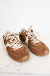 A pair of brown suede sneakers with cream-colored laces and white side stripes, displayed on a light marble surface against a white background.