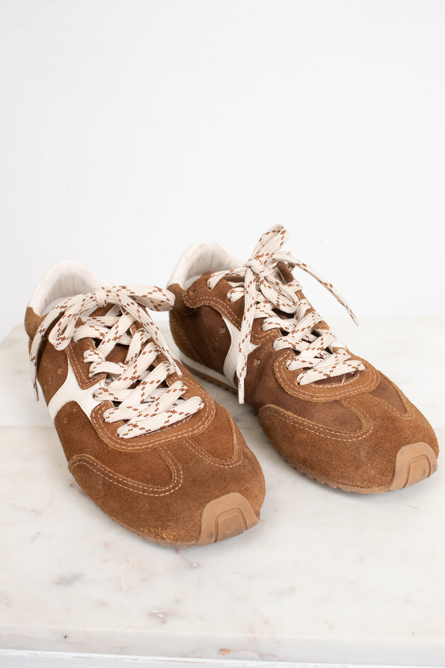 A pair of brown suede sneakers with cream-colored laces and white side stripes, displayed on a light marble surface against a white background.