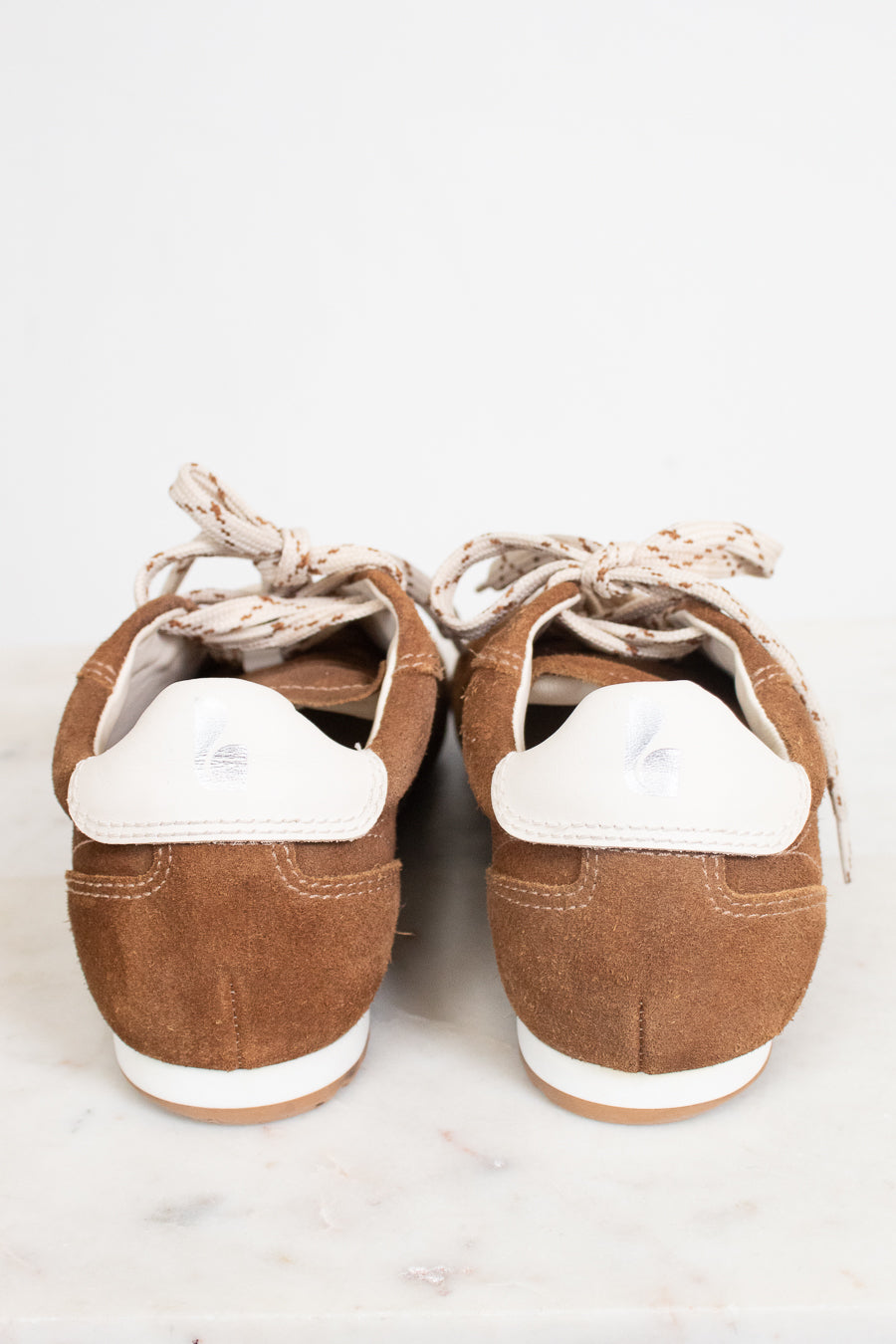 A pair of brown suede low-top sneakers shown from the back, featuring white heel tabs and cream-colored laces, set against a light gray background.