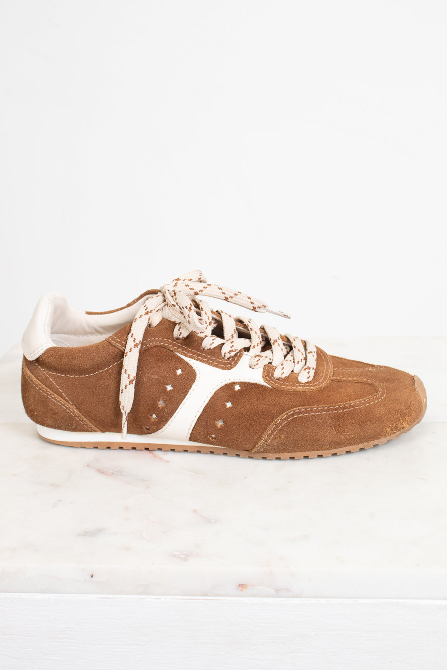 A single brown suede sneaker with cream-colored laces and side stripes, displayed on a white marble surface against a white background.