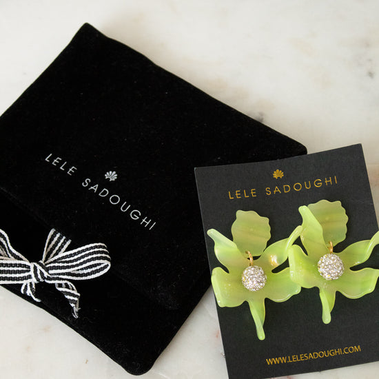 Lele Sadoughi earrings displayed on a black card next to a black velvet jewelry pouch on a light marble surface. The earrings feature bright lime green flower-shaped elements with clear rhinestone centers.