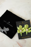 Lele Sadoughi earrings displayed on a black card next to a black velvet jewelry pouch on a light marble surface. The earrings feature bright lime green flower-shaped elements with clear rhinestone centers.