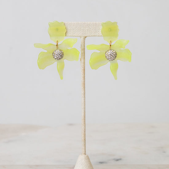 A pair of yellow-green flower-shaped earrings with a textured silver center, displayed on a beige woven jewelry stand against a light gray background.
