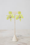 A pair of yellow-green flower-shaped earrings with a textured silver center, displayed on a beige woven jewelry stand against a light gray background.