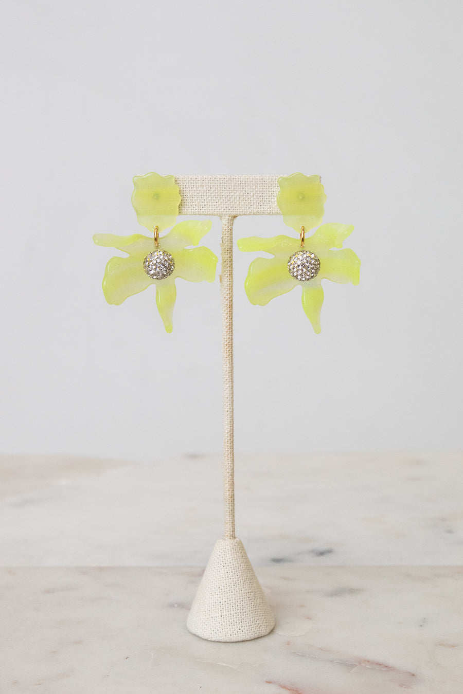 A pair of yellow-green flower-shaped earrings with a textured silver center, displayed on a beige woven jewelry stand against a light gray background.