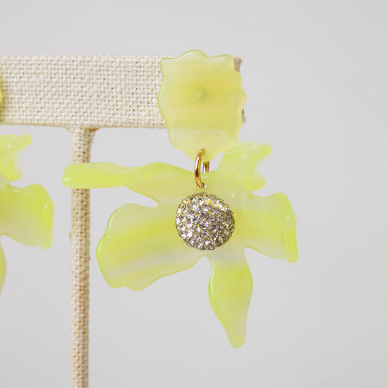 A pair of yellow-green translucent flower-shaped earrings with a gold-tone and crystal-encrusted circular center, displayed on a beige fabric jewelry stand against a light gray background.
