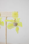 A pair of yellow-green translucent flower-shaped earrings with a gold-tone and crystal-encrusted circular center, displayed on a beige fabric jewelry stand against a light gray background.