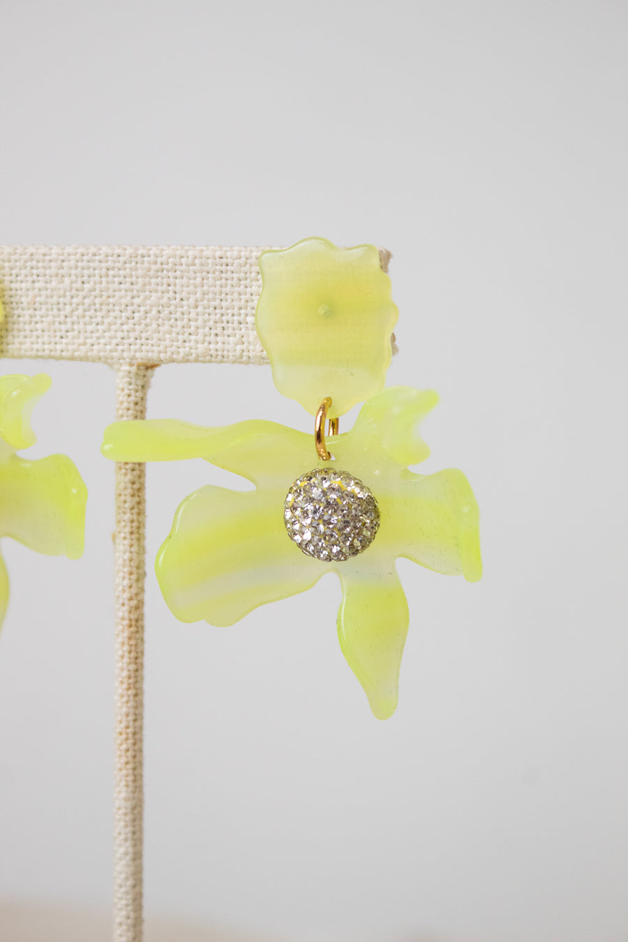A pair of yellow-green translucent flower-shaped earrings with a gold-tone and crystal-encrusted circular center, displayed on a beige fabric jewelry stand against a light gray background.