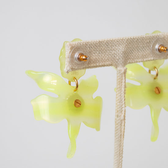 Yellow-green translucent flower-shaped dangle earrings with small gold-tone details, displayed on a beige fabric jewelry stand against a light gray background.