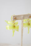 Yellow-green translucent flower-shaped dangle earrings with small gold-tone details, displayed on a beige fabric jewelry stand against a light gray background.